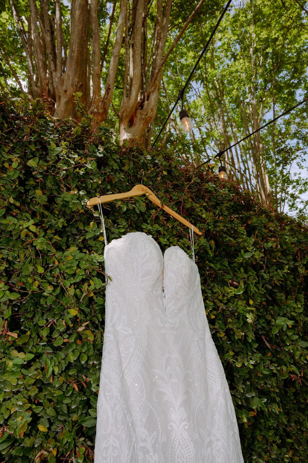 White lace wedding dress hanging on a wooden hanger outdoors against a lush green hedge and trees.