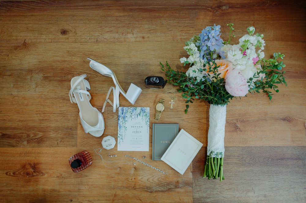 Elegant wedding flat lay with bridal heels, floral bouquet, invitations, and accessories on wood floor.