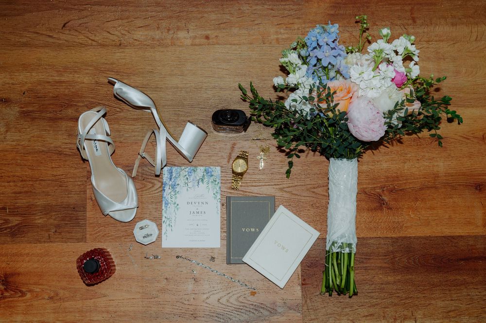 Wedding flat lay featuring bridal bouquet, heels, invitations, rings, and accessories on wood floor.