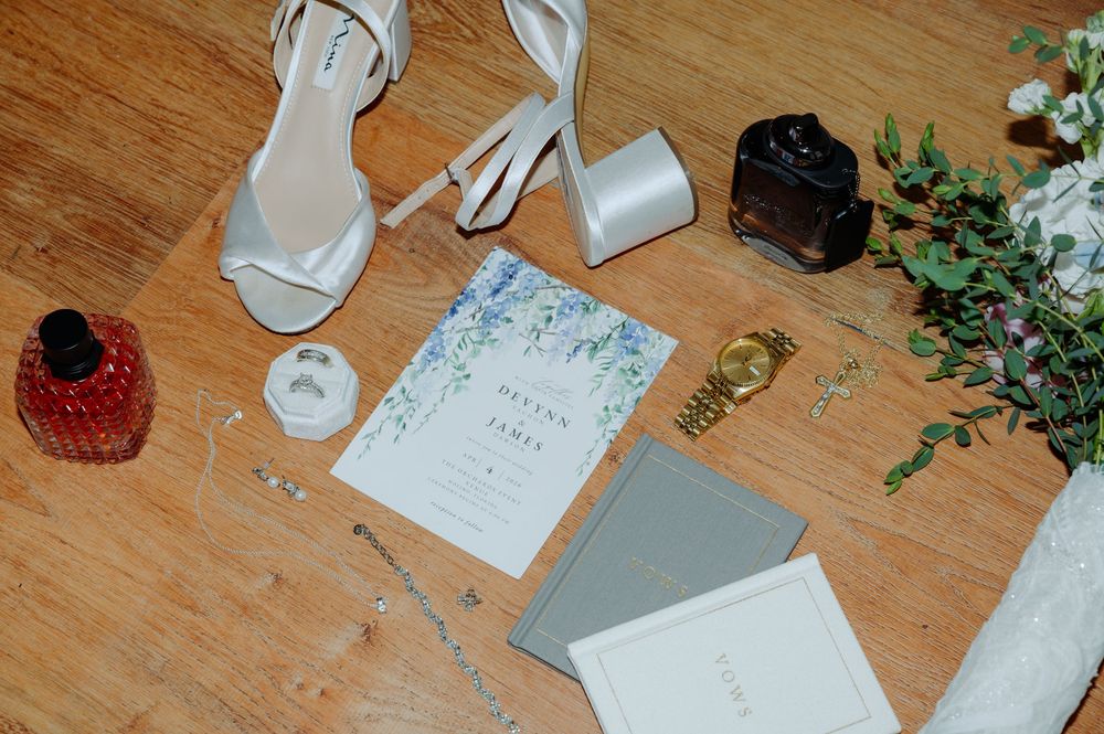 Elegant wedding flat lay with white heels, floral invitation, perfume bottles, and greenery on wood floor.