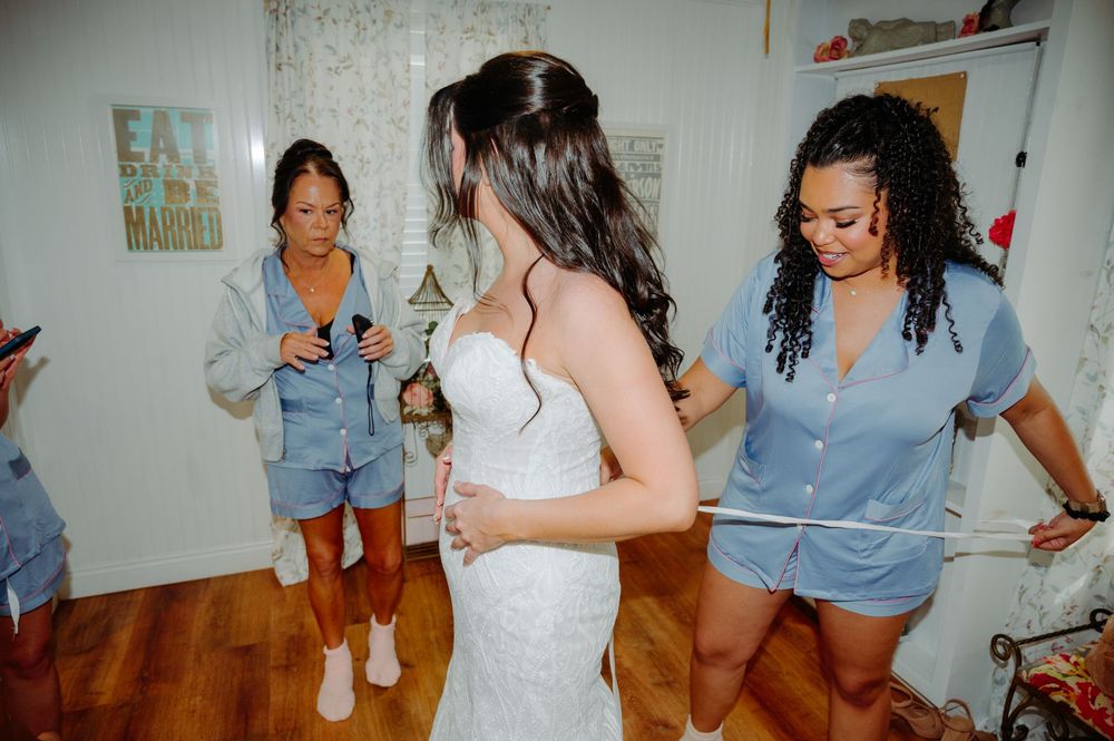 Bridesmaids in blue outfits helping bride into white lace wedding gown in a cozy bridal suite.