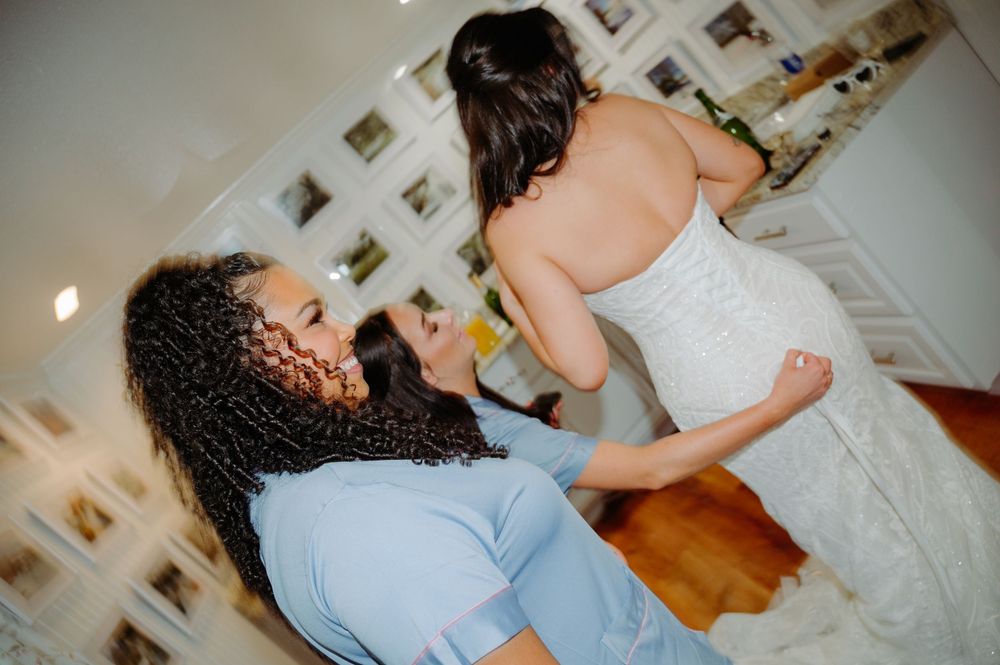 Bridesmaid helping bride with her wedding dress during bridal preparations indoors.