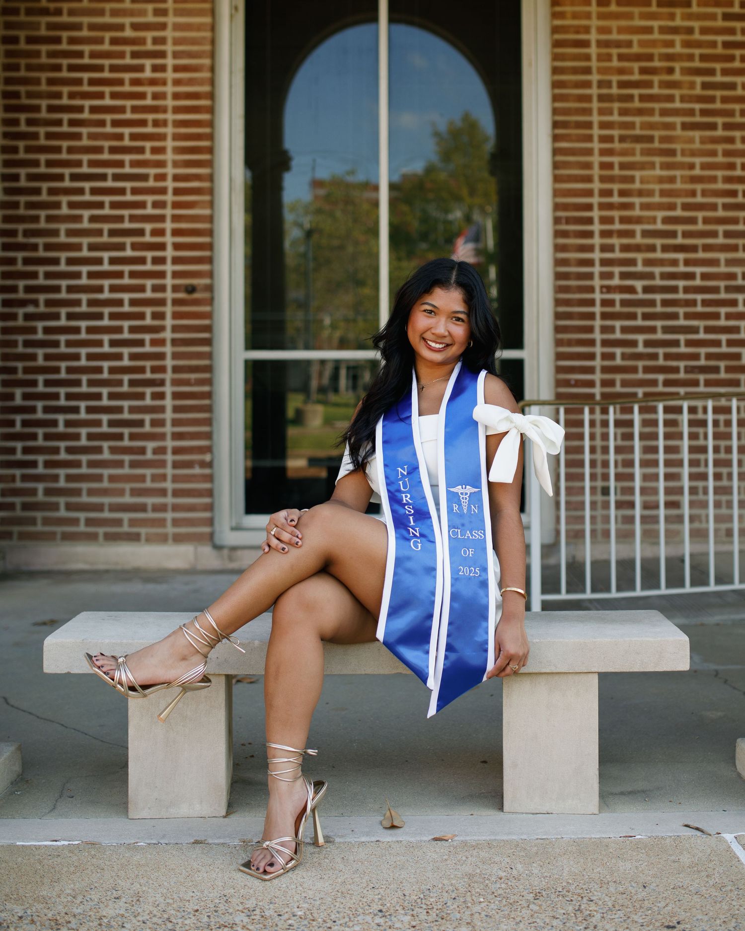 Young woman in white dress and blue graduation sash sits on stone bench outside brick building, smiling.