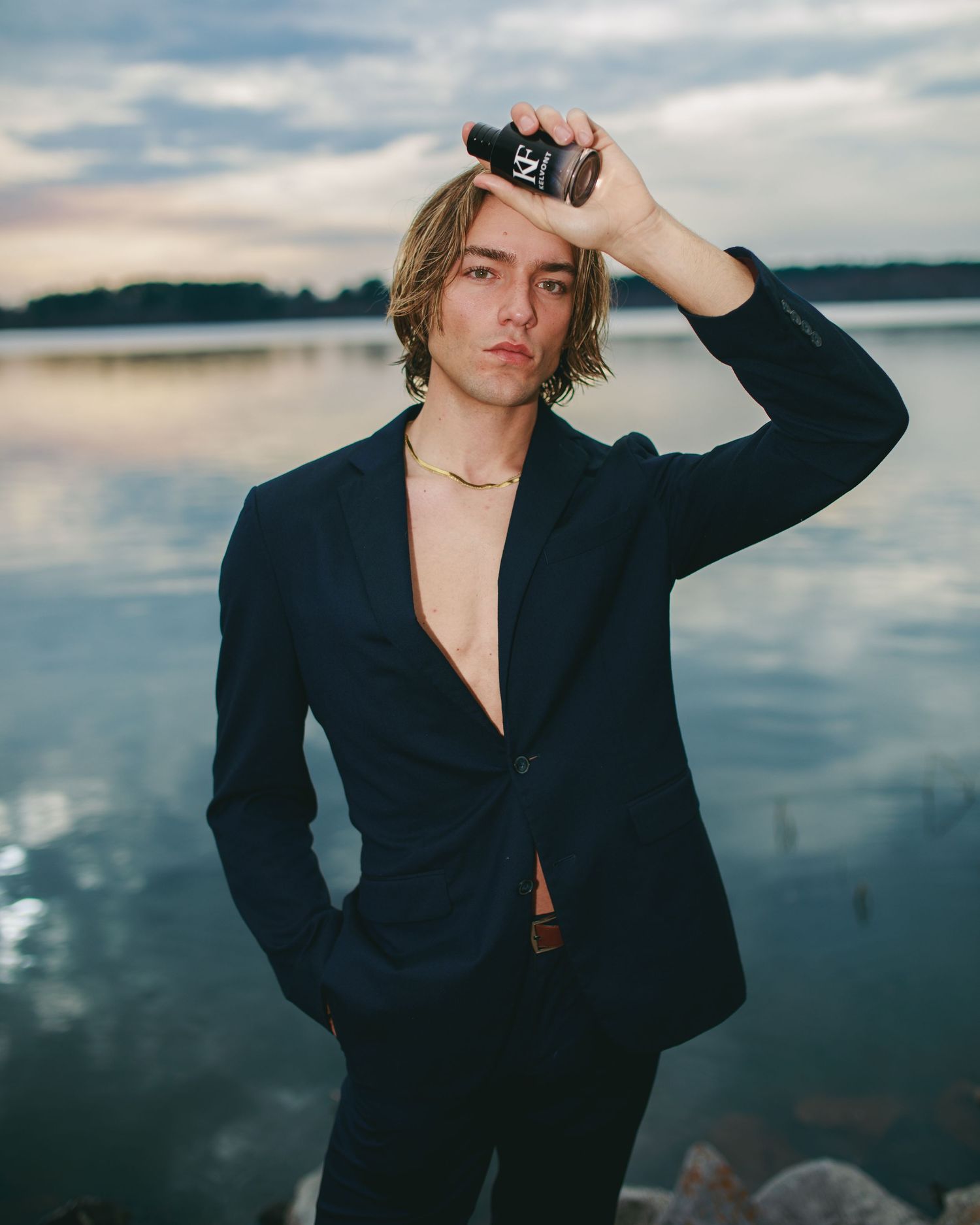 Young man in navy blazer holding a dark perfume bottle by a rocky lakeside at dusk.