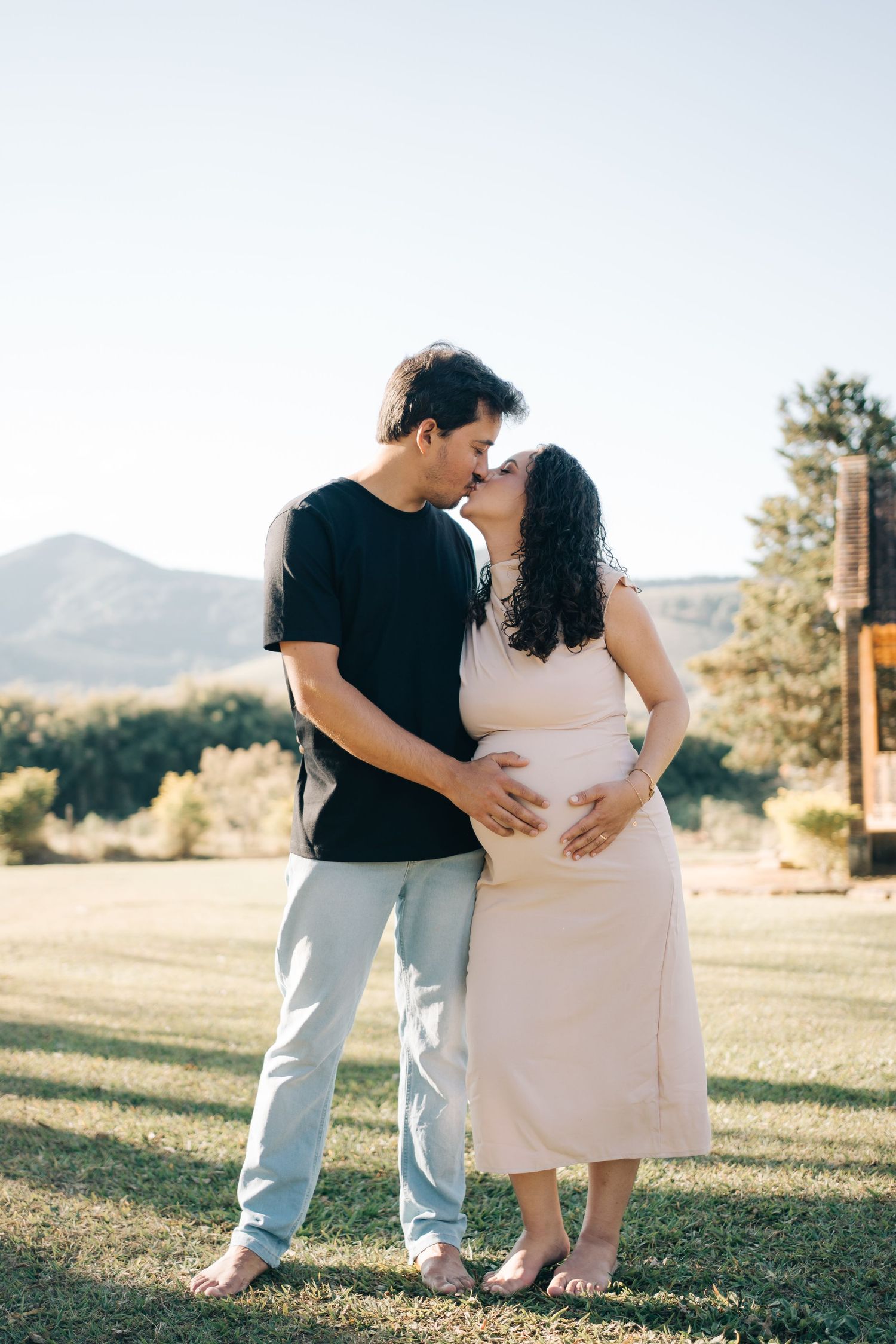 Liliana e Bruno se beijam com as mãos sobre a barriga, diante das montanhas na Fazenda Vintém, em Santa Rita do Sapucaí.