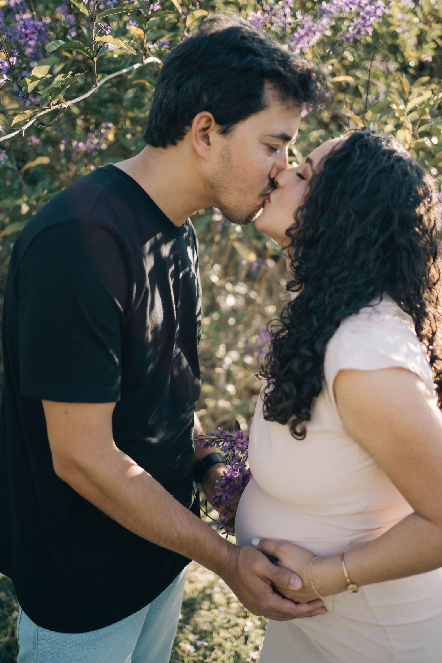 Liliana e Bruno se beijam diante das flores, com as mãos sobre a barriga, durante o ensaio de gestante na Fazenda Vintém.