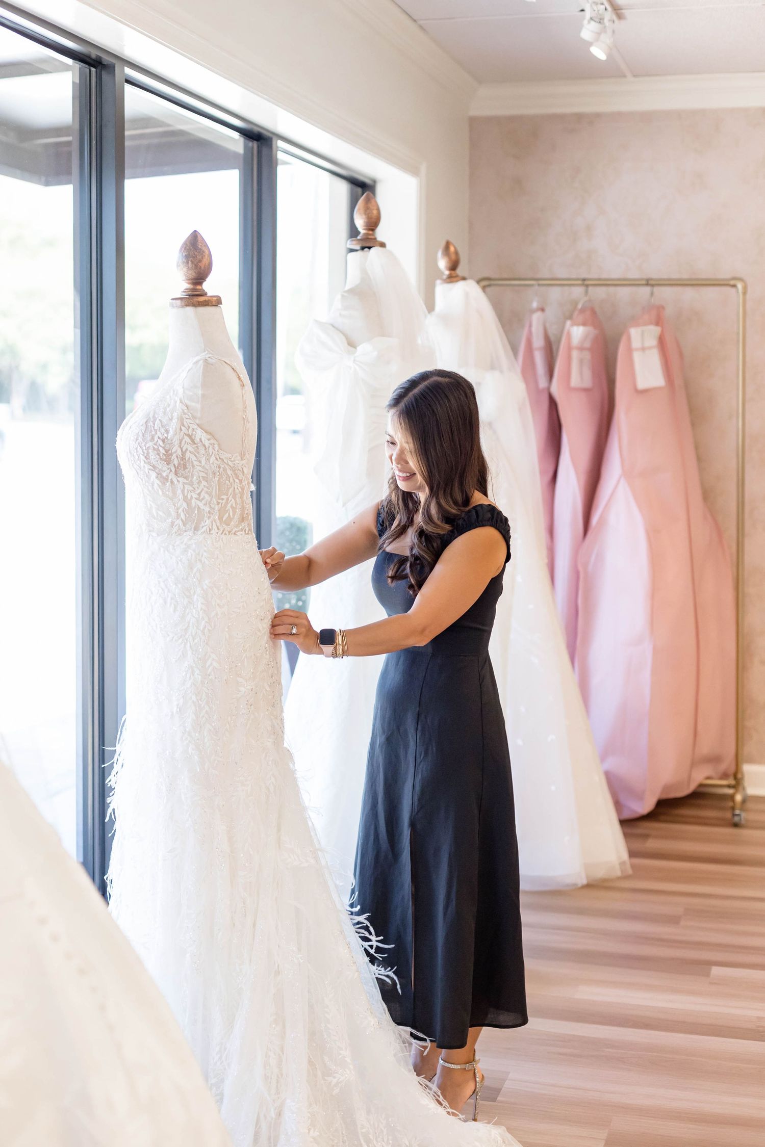A woman in a navy dress adjusts a white lace wedding gown on a mannequin in a bright bridal boutique.