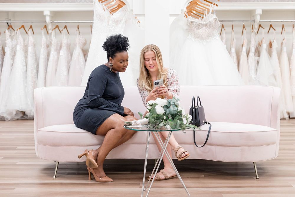 Two women sit on a pink sofa in a bridal boutique, looking at a phone surrounded by wedding dresses.