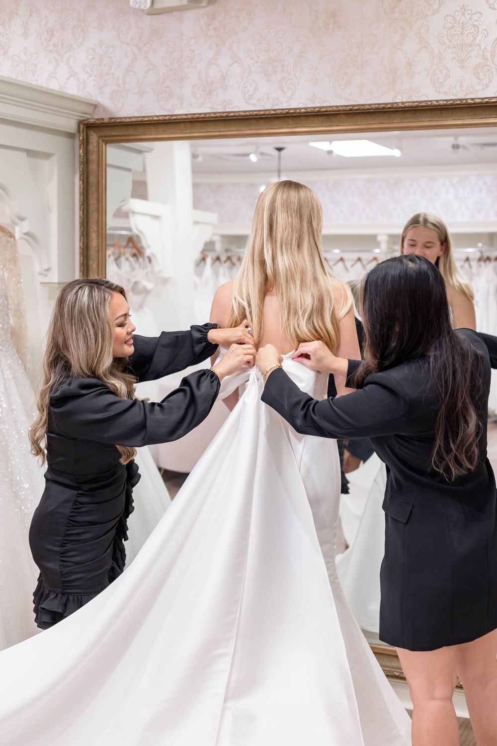 Two bridal consultants in black dresses fasten the back of a white wedding gown on a blonde bride in a bridal boutique.