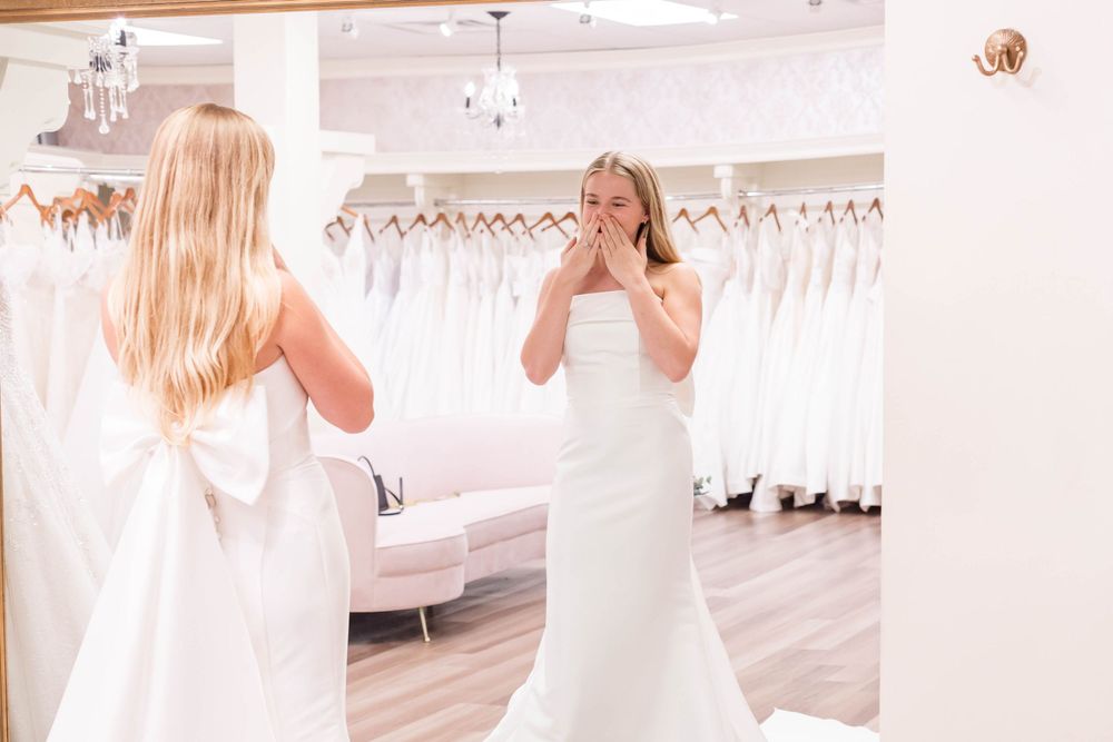 Bride trying on a white wedding dress in a bridal boutique, covering her mouth in an emotional reaction before a mirror.