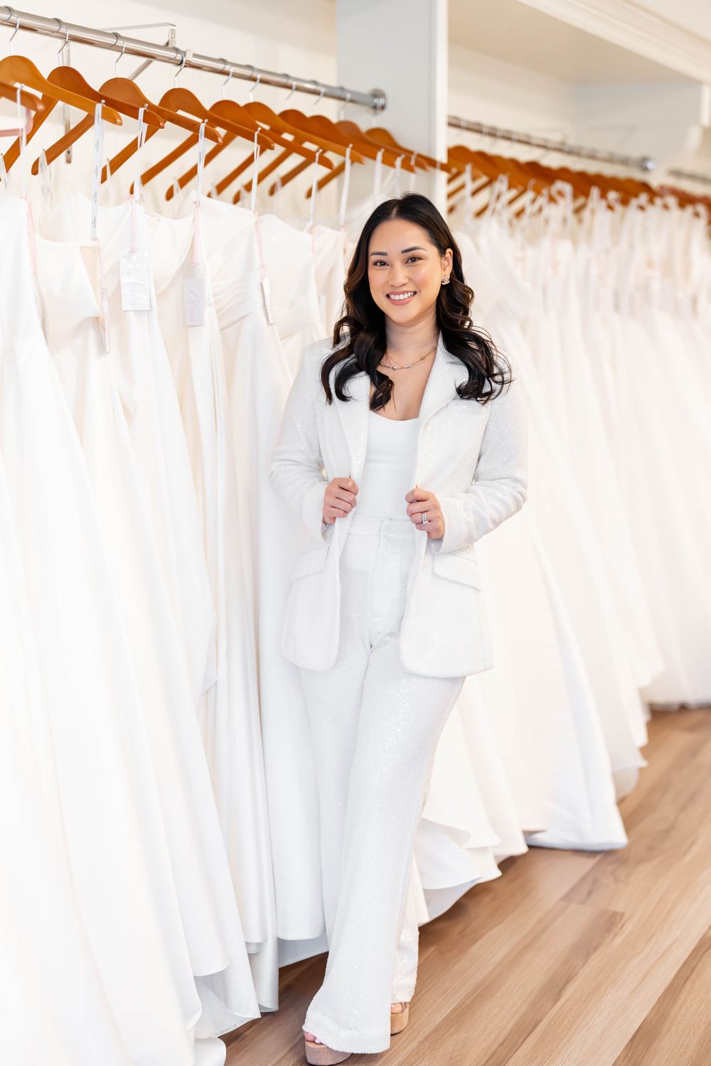 Smiling woman in white pantsuit poses among elegant bridal gowns on hangers in a wedding dress boutique.