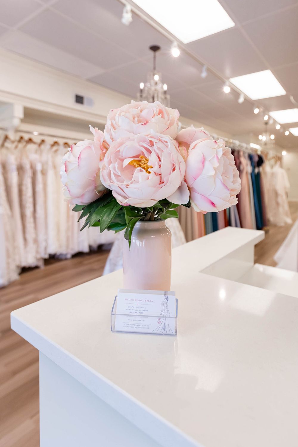 Pink peony bouquet in white vase on bridal boutique counter with wedding dresses displayed in background.