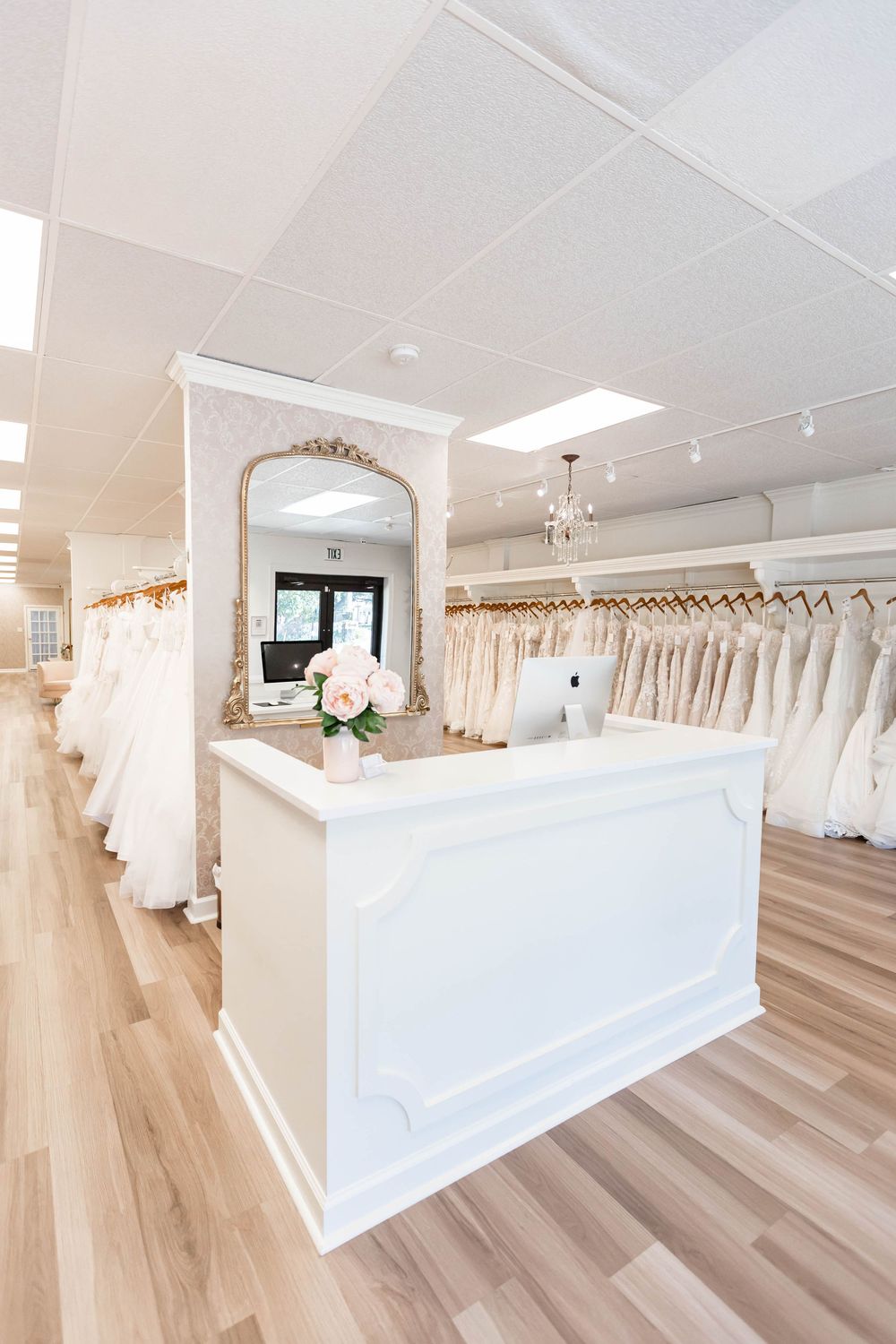 Elegant bridal boutique interior with white reception desk, ornate mirror, and rows of wedding gowns on display racks.