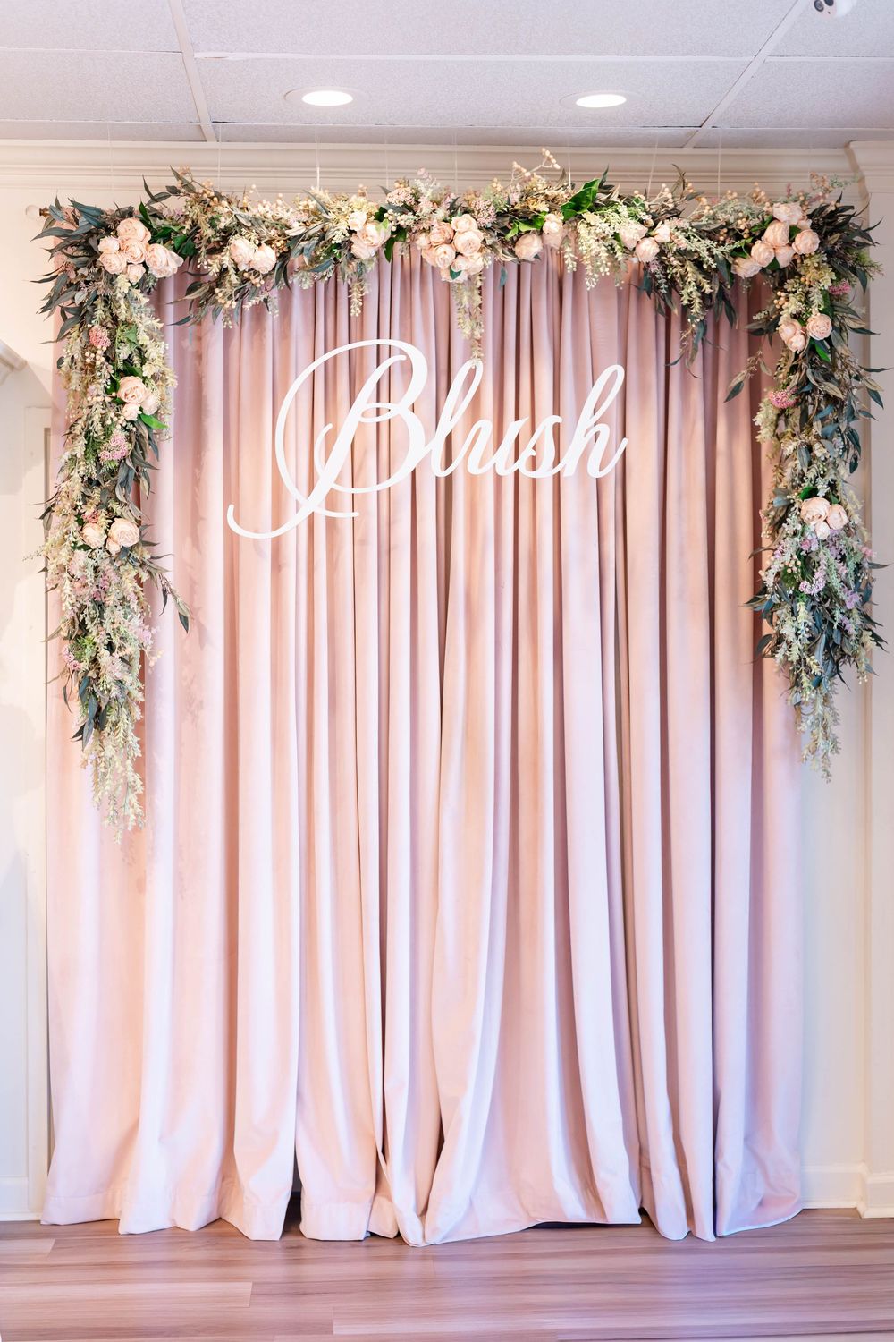 Elegant blush pink draped backdrop with floral arch and white script Blush sign for wedding or event decor.
