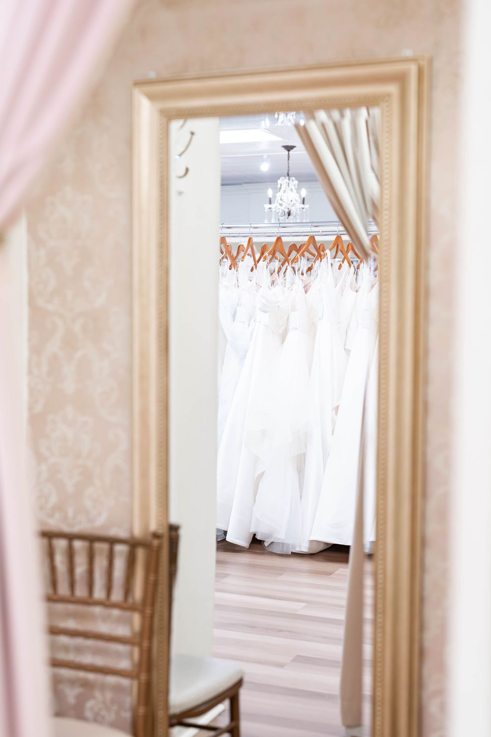 Gold-framed mirror reflecting a bridal boutique showroom with white wedding dresses on hangers and an elegant chandelier.