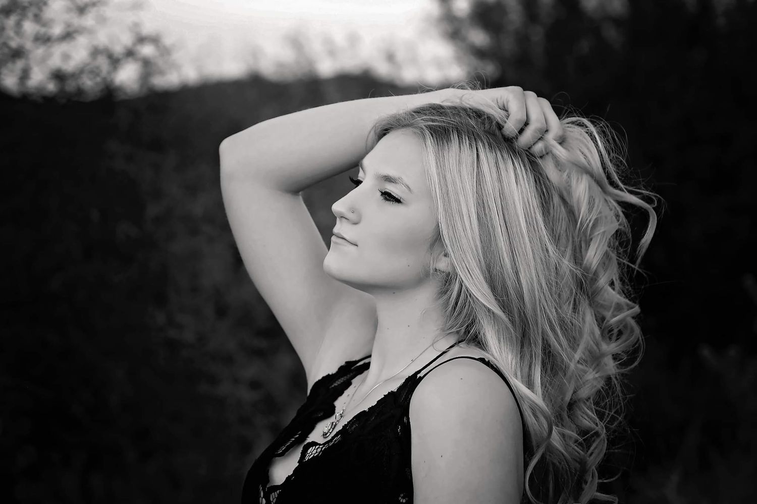 Black and white portrait of a woman with long wavy hair, one hand raised to her head, gazing off to the side outdoors.