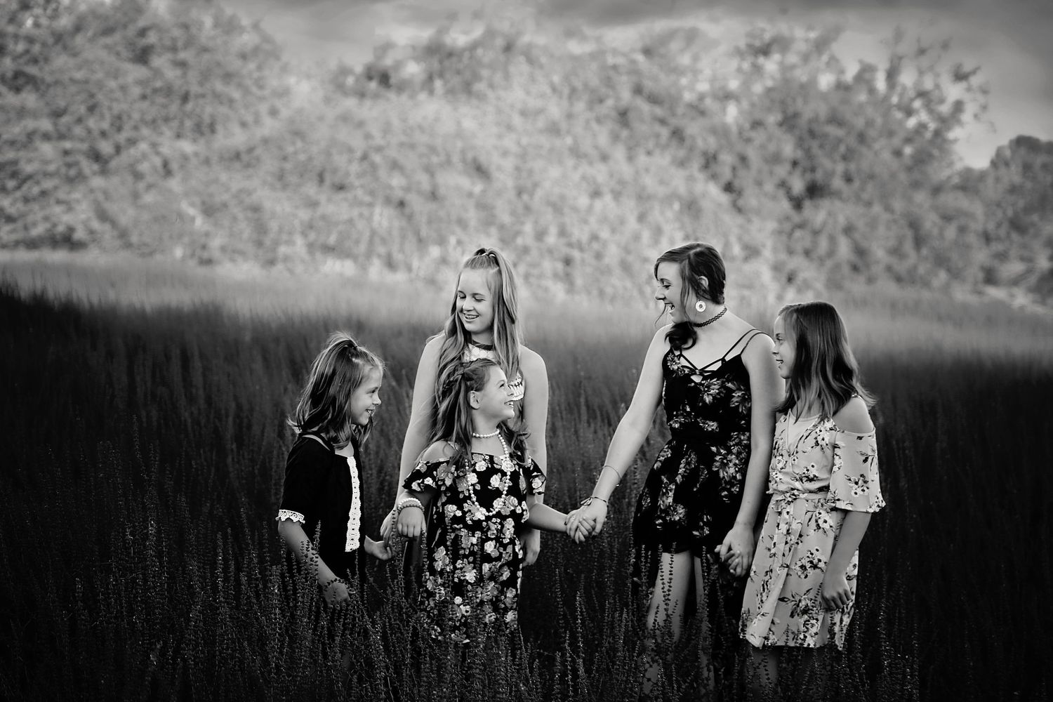 Black and white photo of four girls and a woman holding hands while walking through tall grass in a field.
