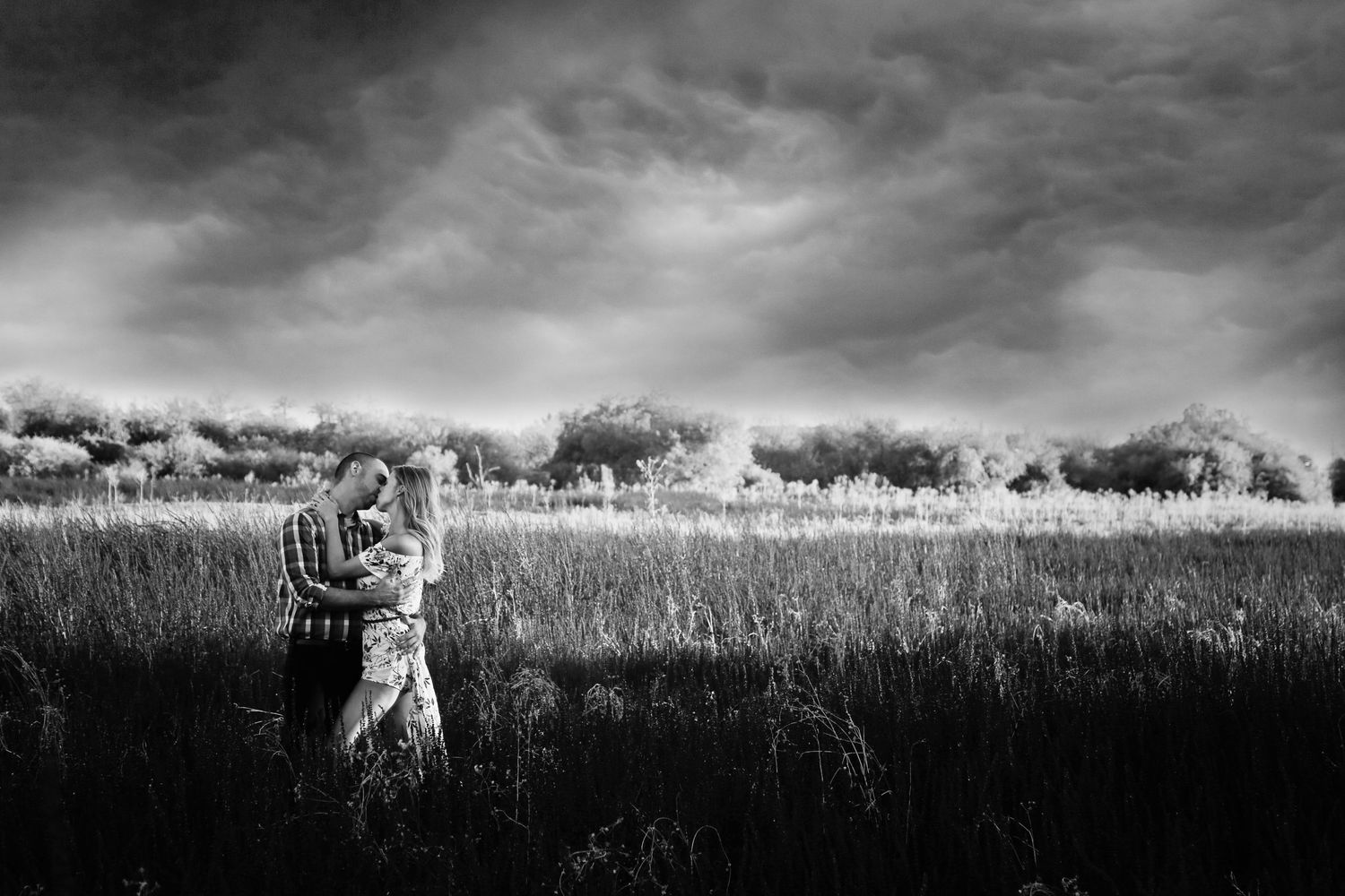 Black and white photo of a couple embracing in a grassy field beneath a dramatic stormy sky.