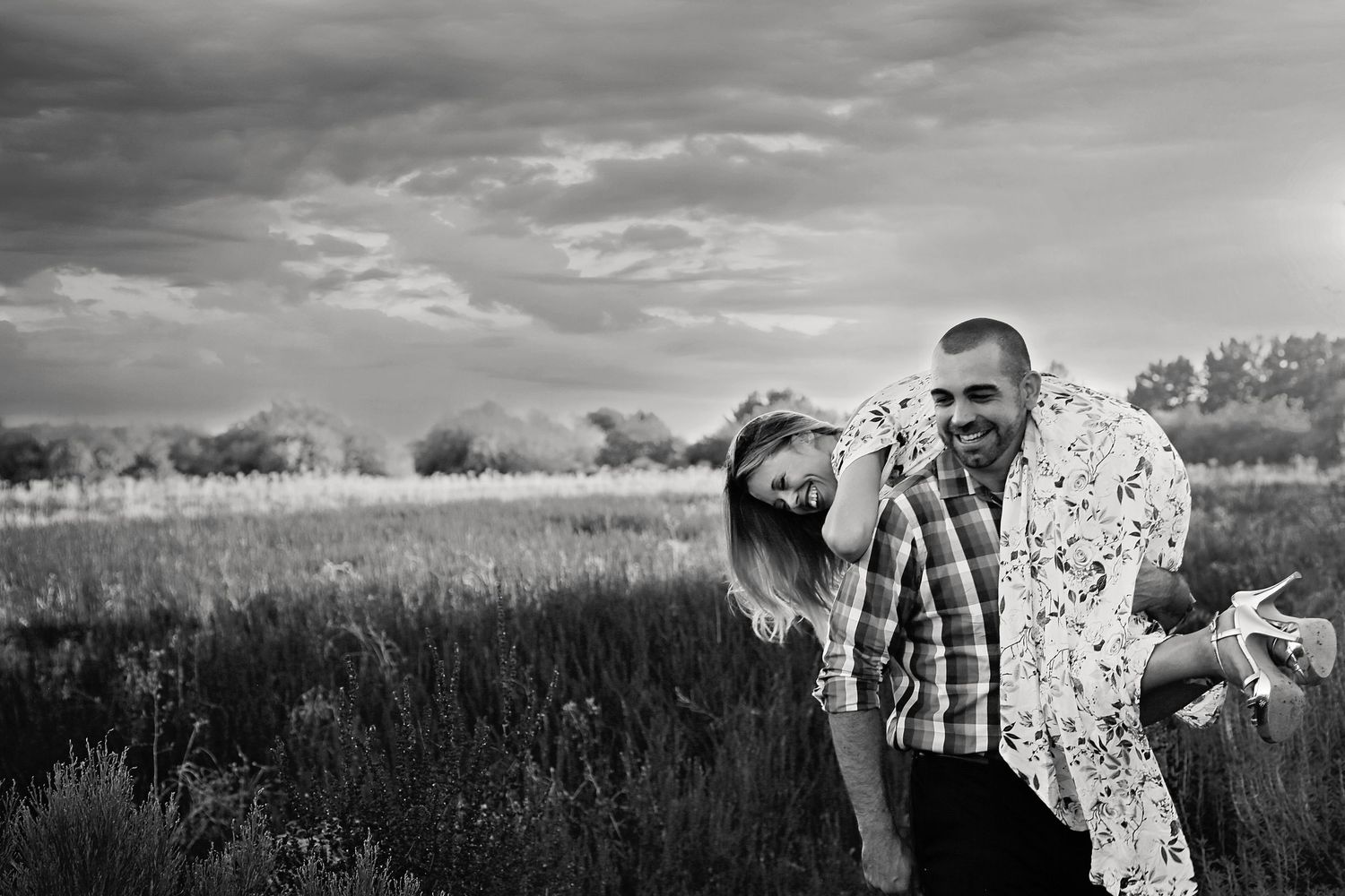 Black and white photo of a smiling man carrying a laughing woman over his shoulder in a grassy field under stormy skies.