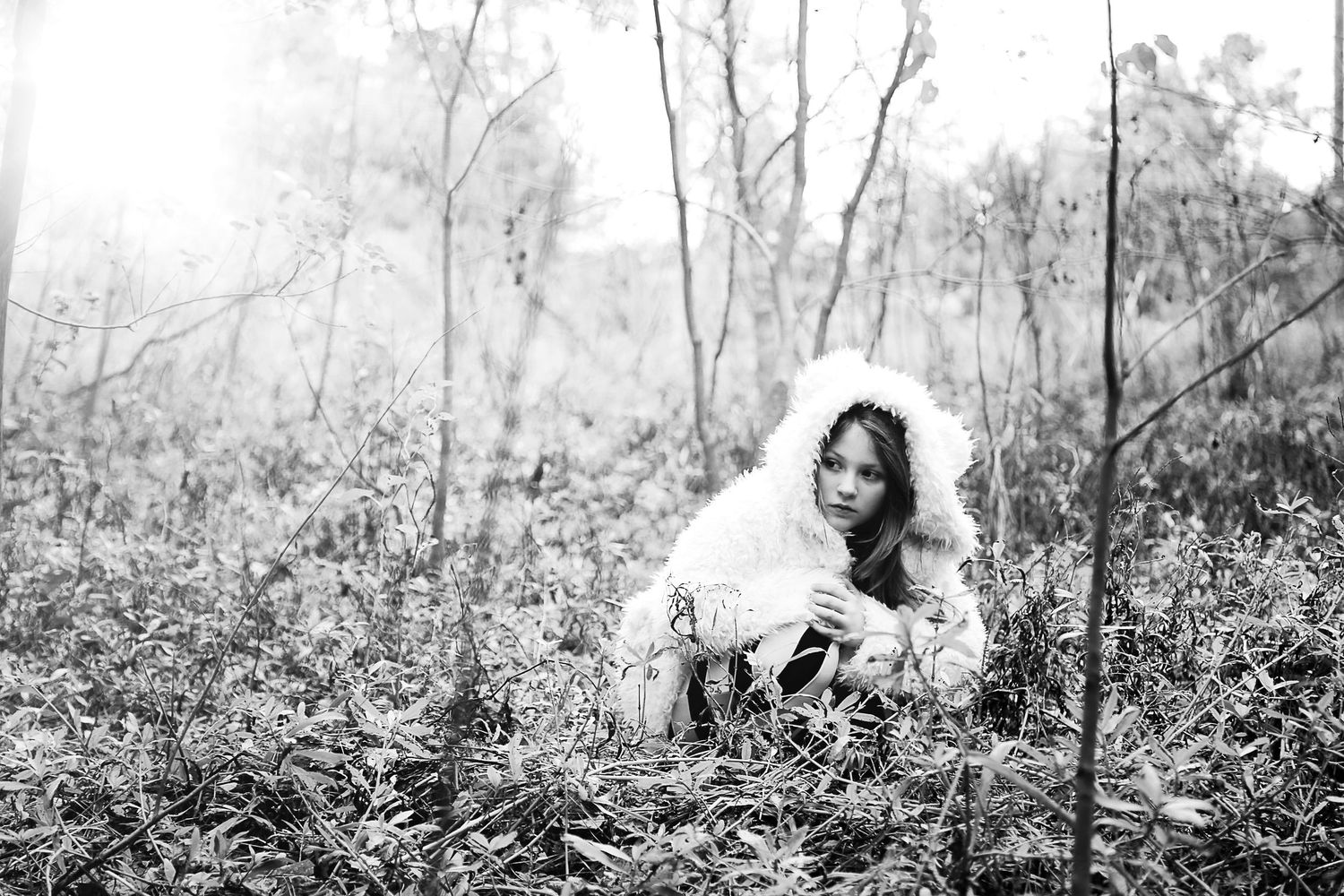 Black and white portrait of a girl in a fluffy white hooded coat sitting among bare trees in a forest.