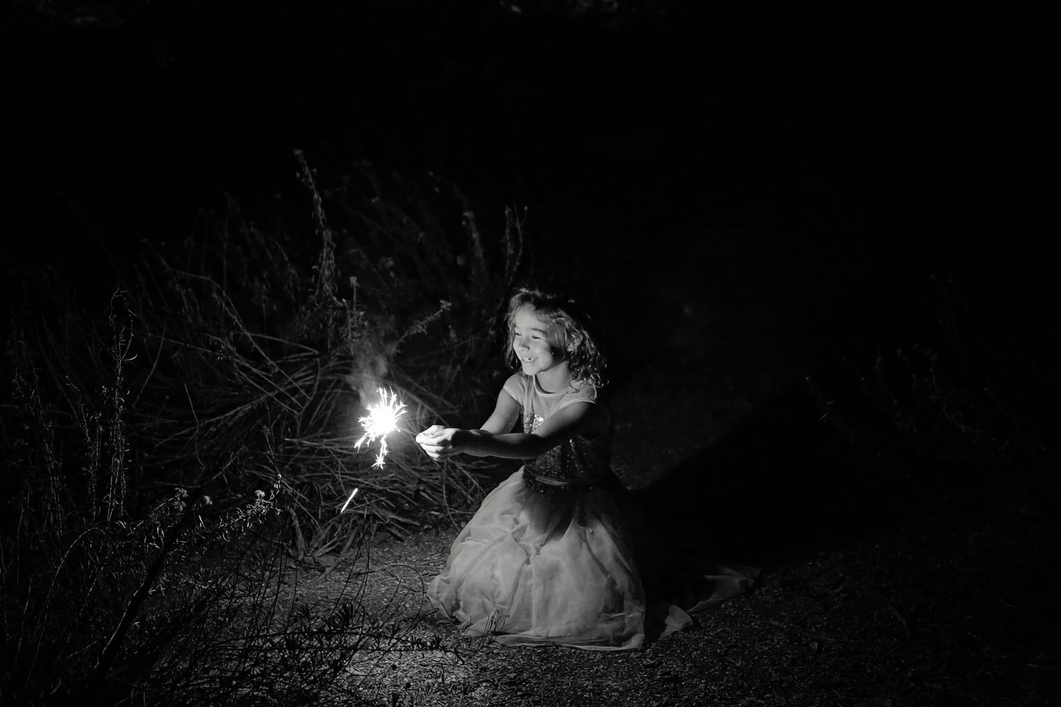 Black and white photo of a smiling girl in a tulle dress kneeling in the dark and holding a glowing sparkler.