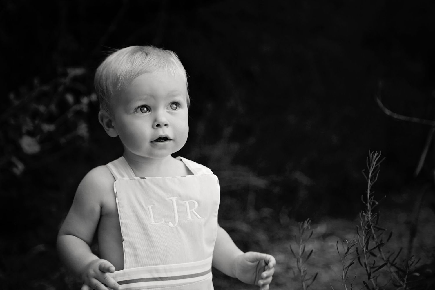 Black and white portrait of a toddler in a monogrammed romper looking curiously upward against a dark background.