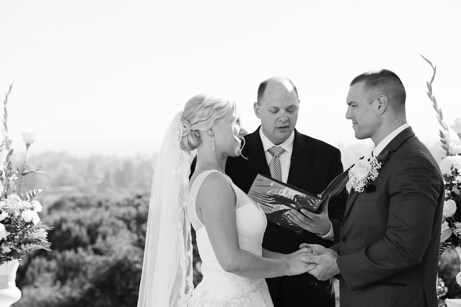 Black and white wedding ceremony with a bride and groom holding hands as an officiant reads before them outdoors.