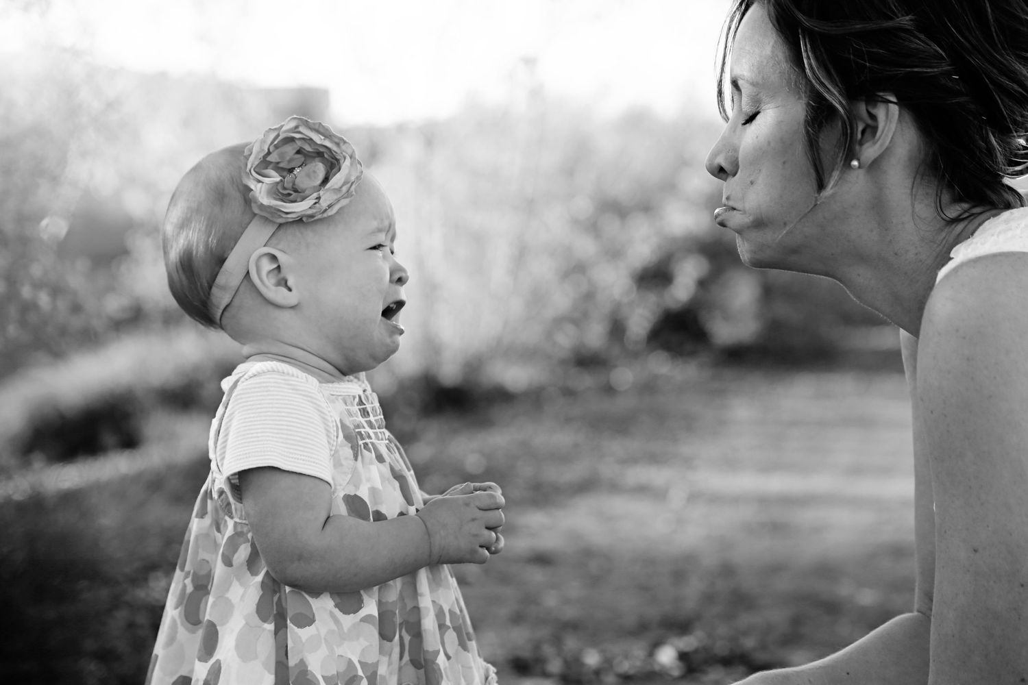 Black and white photo of a mother blowing a kiss to a crying toddler girl wearing a floral headband outdoors.