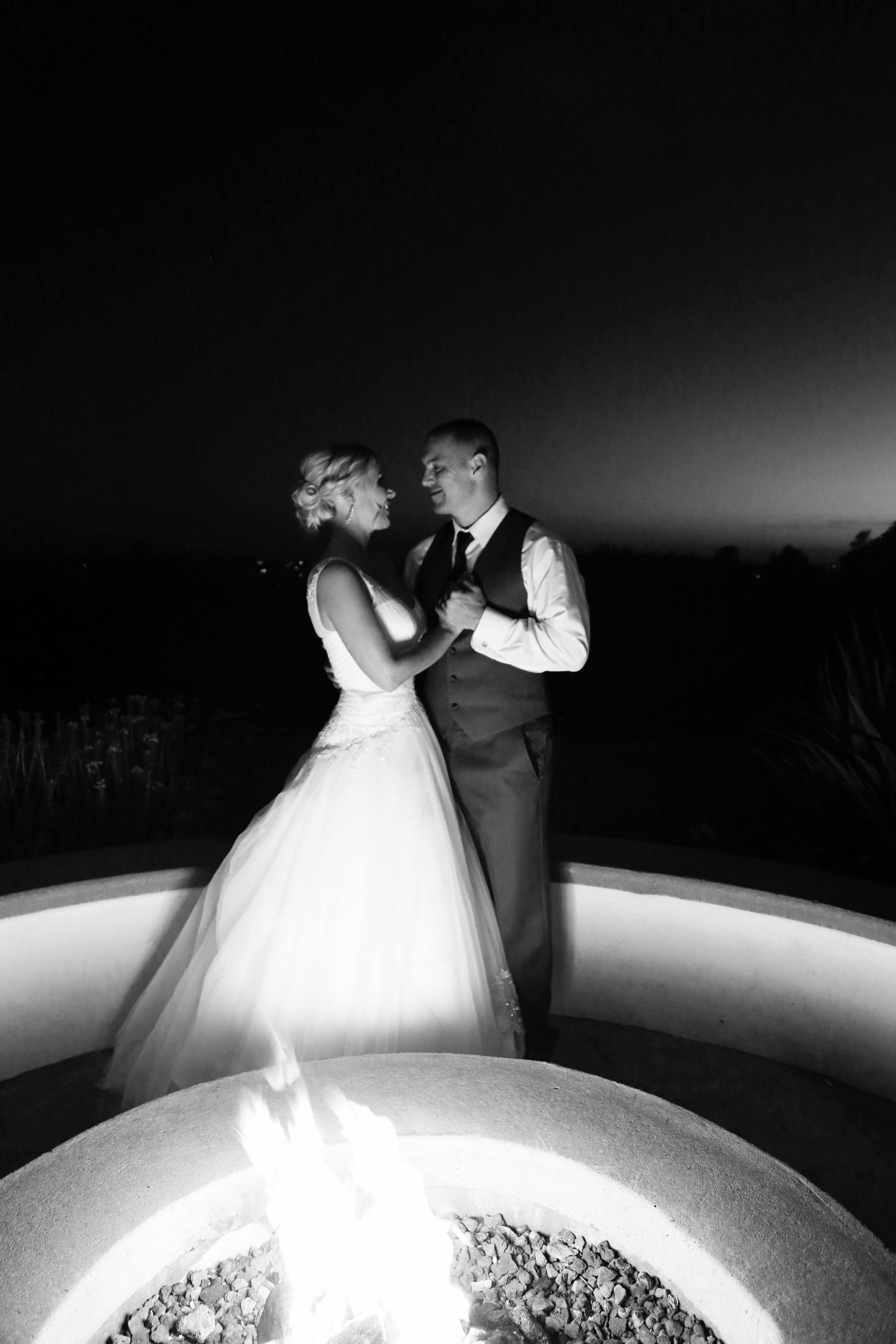 Black and white photo of a bride and groom dancing together beside a glowing fire pit at night.