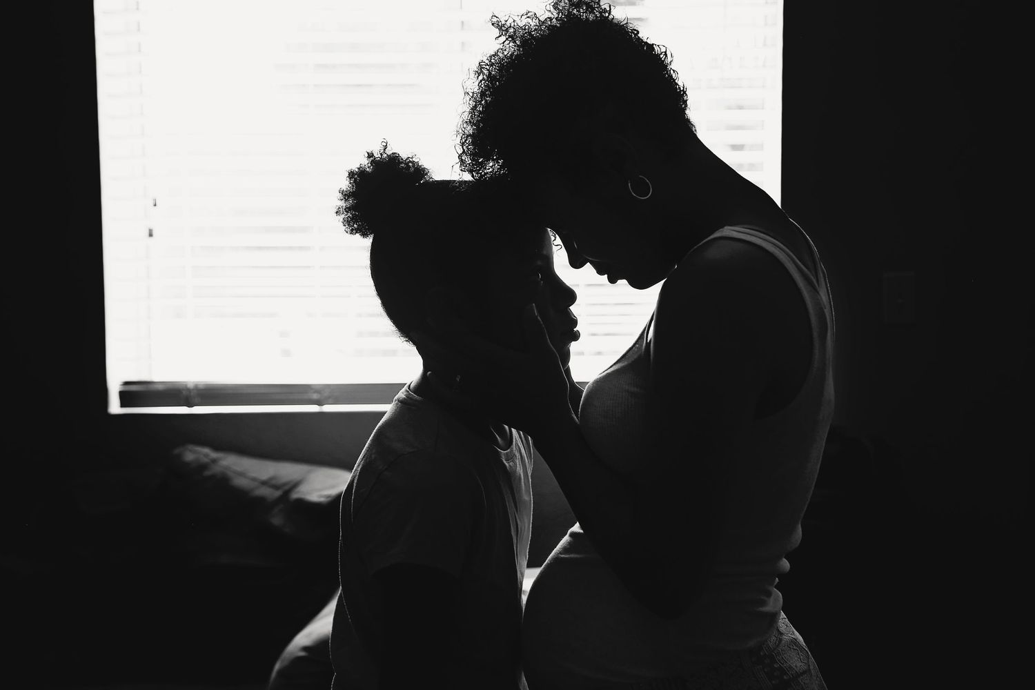 Black and white silhouette of a pregnant mother touching foreheads with her child in front of a bright window.