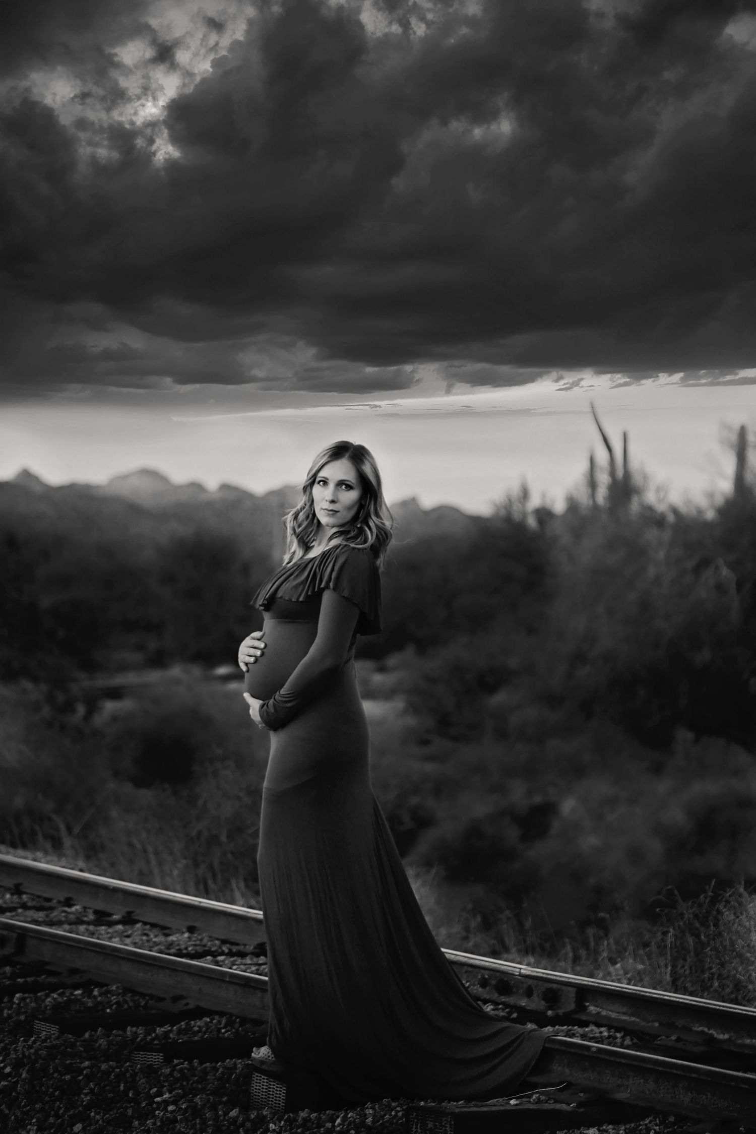 Black and white portrait of a pregnant woman in a flowing gown standing on railroad tracks beneath a stormy desert sky.