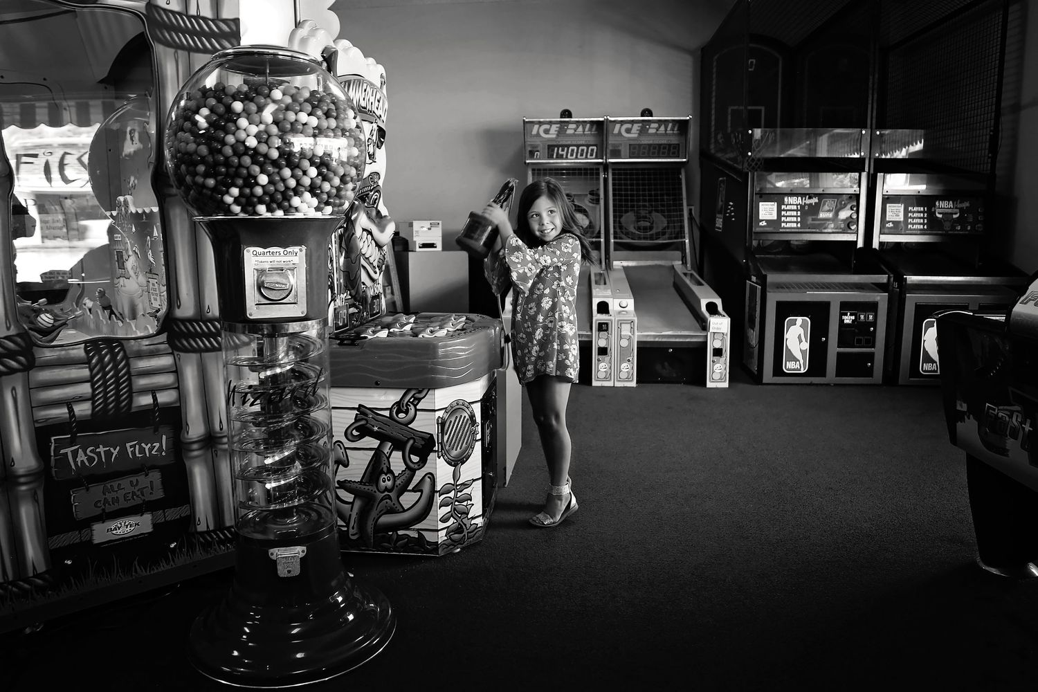 Black and white photo of a smiling girl in a patterned dress posing beside a gumball machine in a retro arcade.