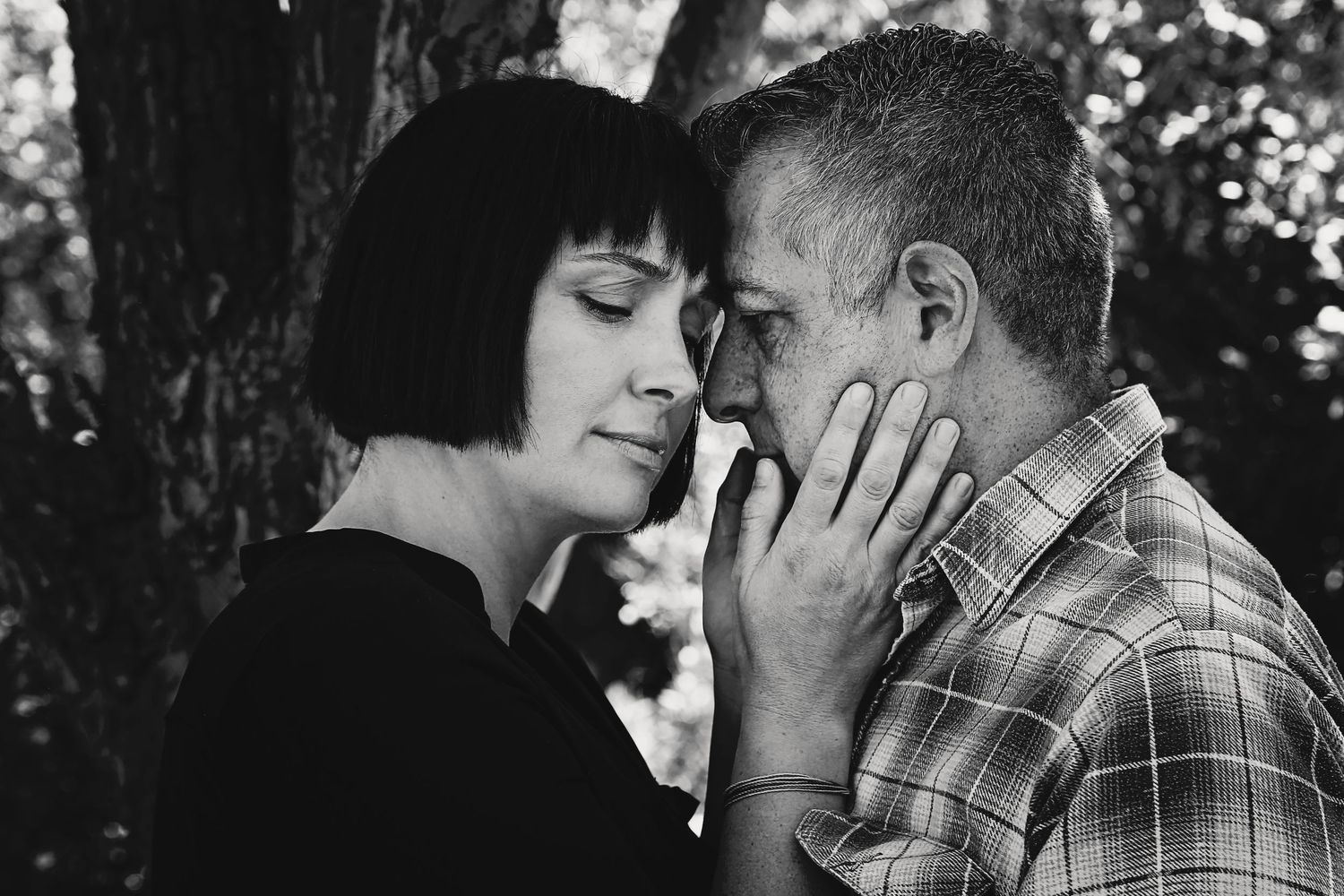 Black and white close-up of a couple sharing a tender moment, foreheads touching, beneath trees outdoors.