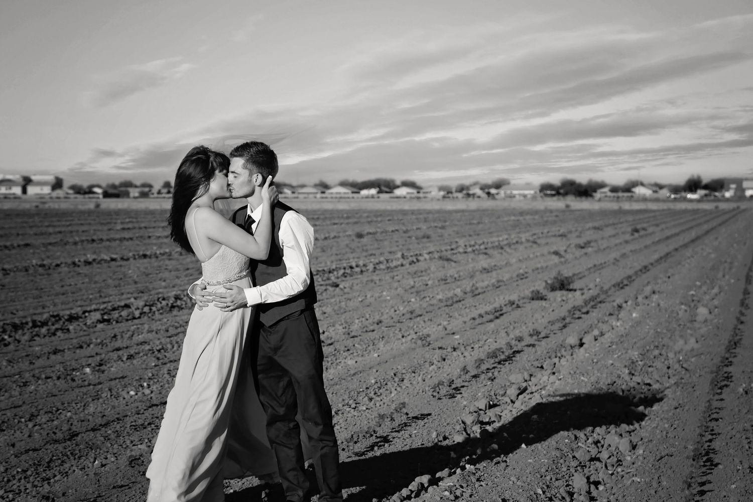 Black and white photo of a couple kissing in a plowed field beneath a wide cloudy sky with distant houses.