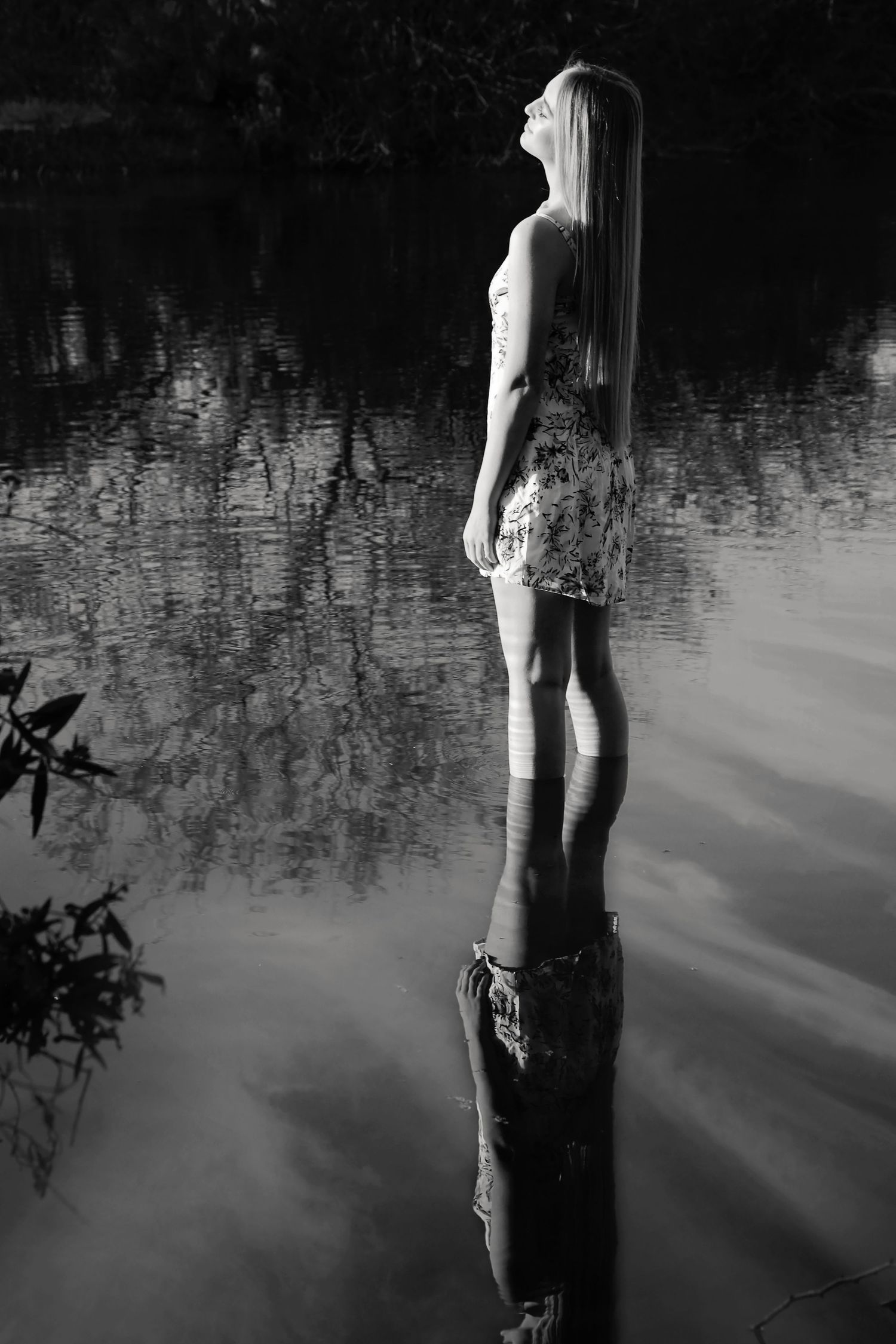 Black and white photo of a woman in a floral dress standing in shallow water, her reflection mirrored on the lake surface.