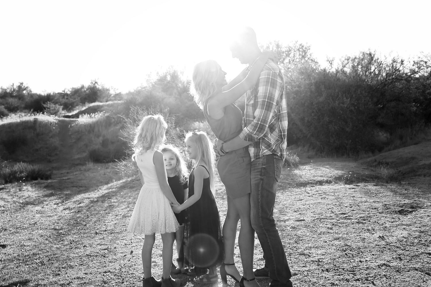 Black and white photo of a family with young girls embracing outdoors in a sunlit desert landscape.