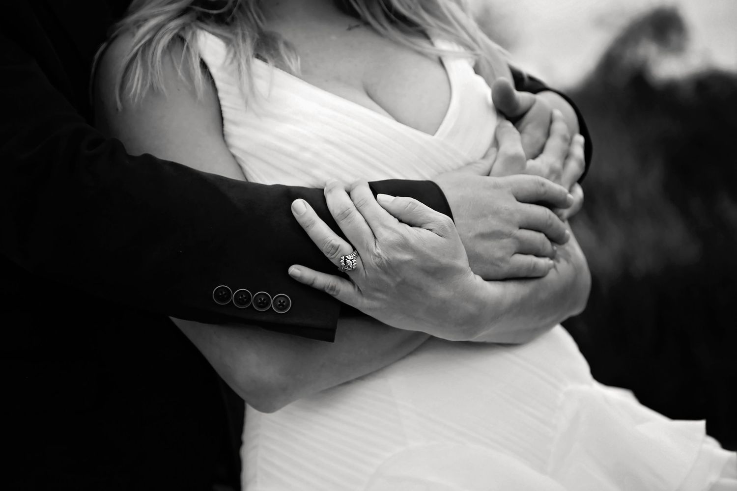 Black and white close-up of a groom's arms embracing a bride in a white gown, her ring visible on her hand.
