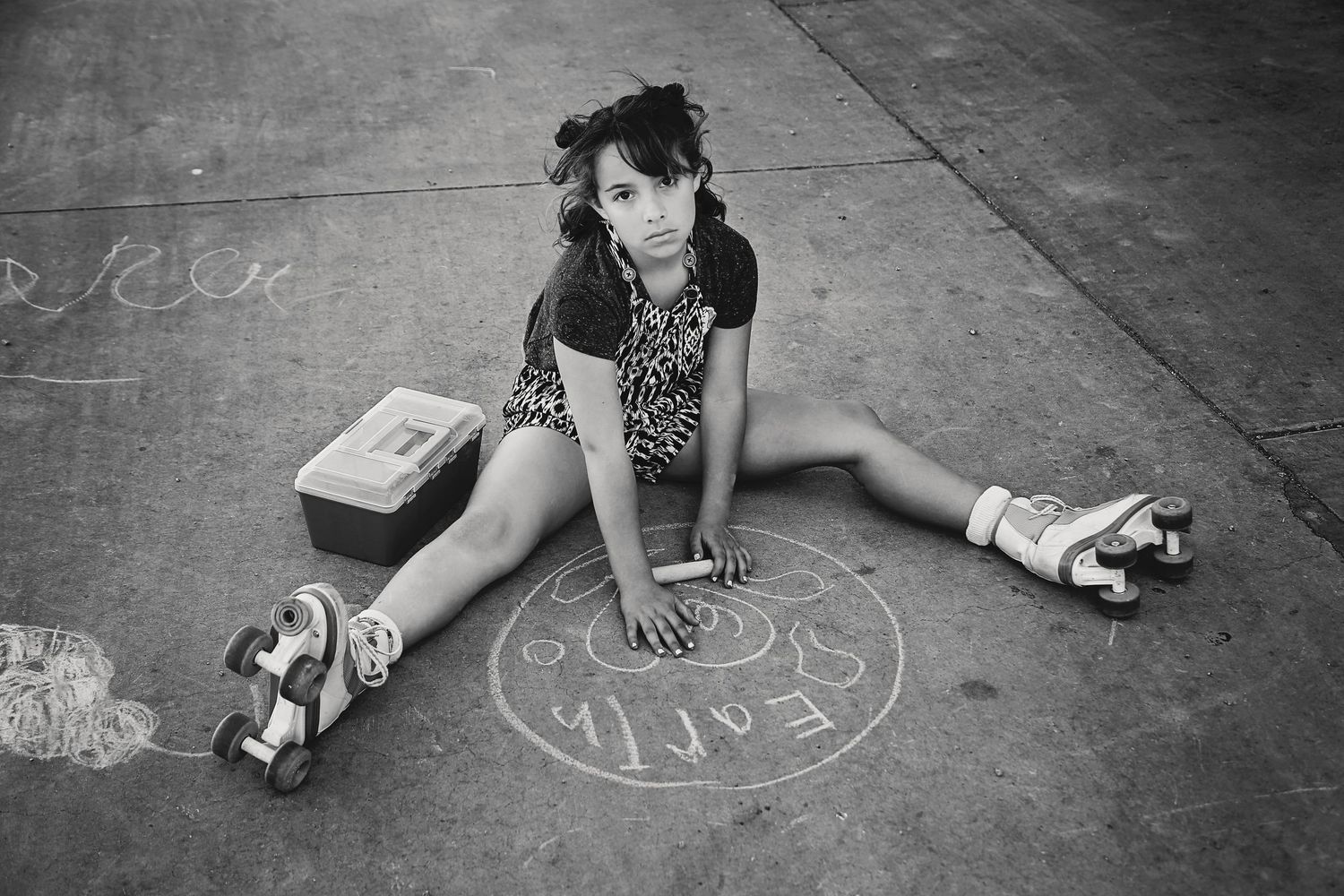 Black and white photo of a girl in roller skates sitting on pavement beside a small case and a chalk drawing.