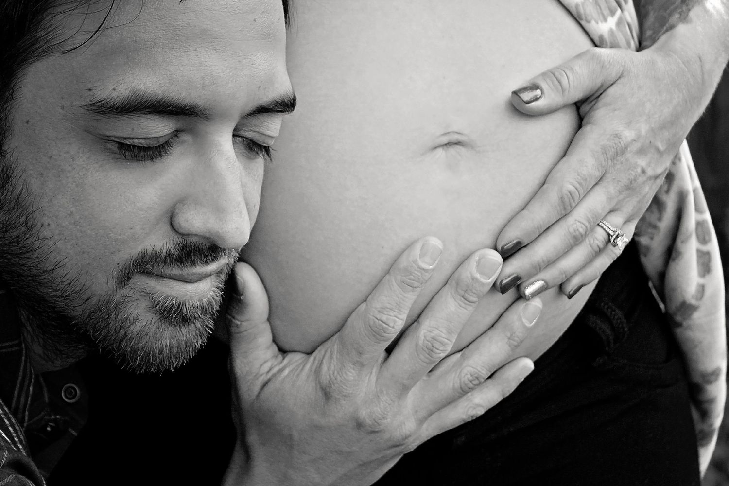 Black and white close-up of a bearded man resting his face against a pregnant belly as hands cradle it.