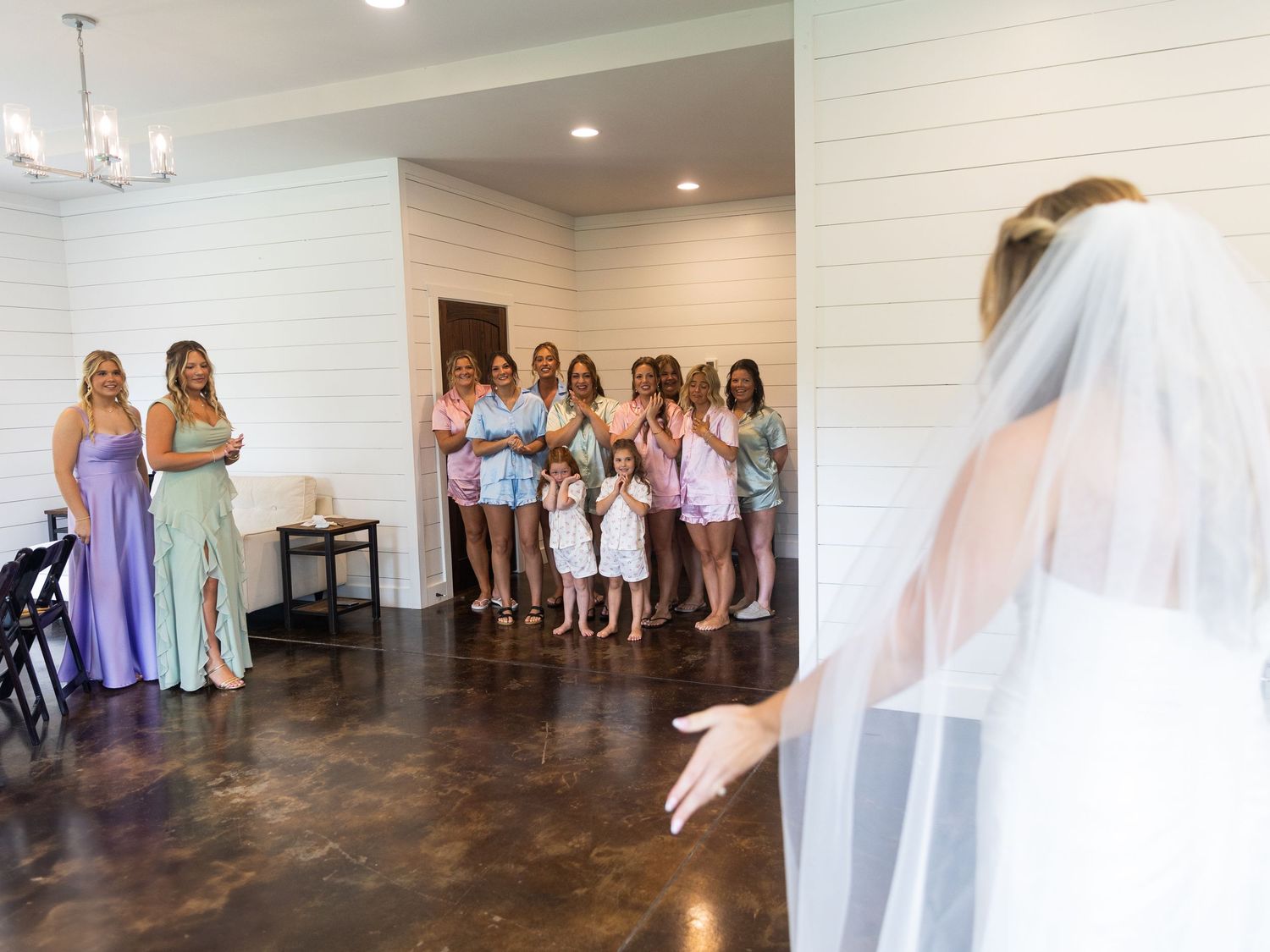 Bride in white gown and veil reveals her wedding look to bridesmaids and guests in a decorated venue.