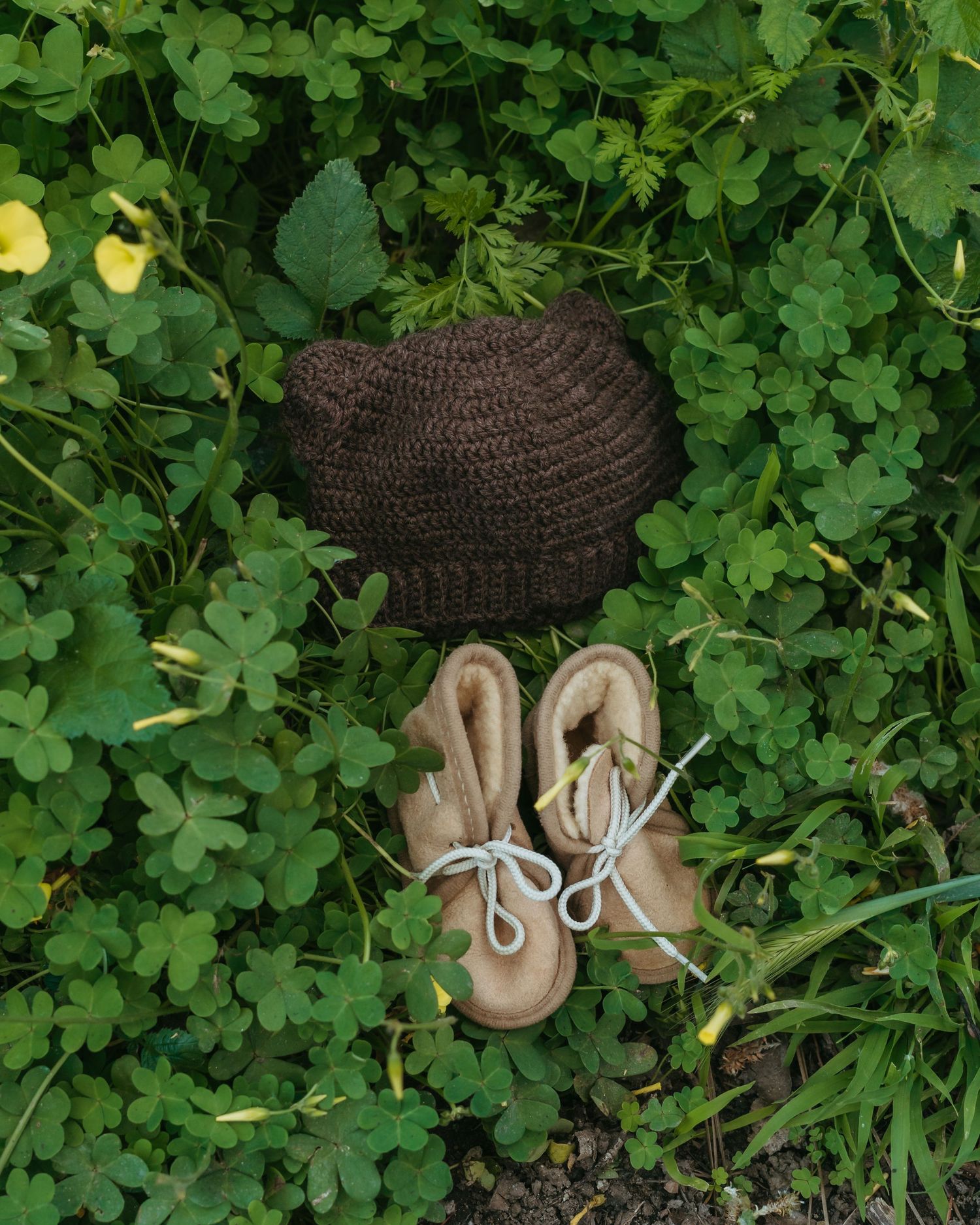 Brown crochet baby hat and tan leather booties with white laces placed on lush green clover grass.