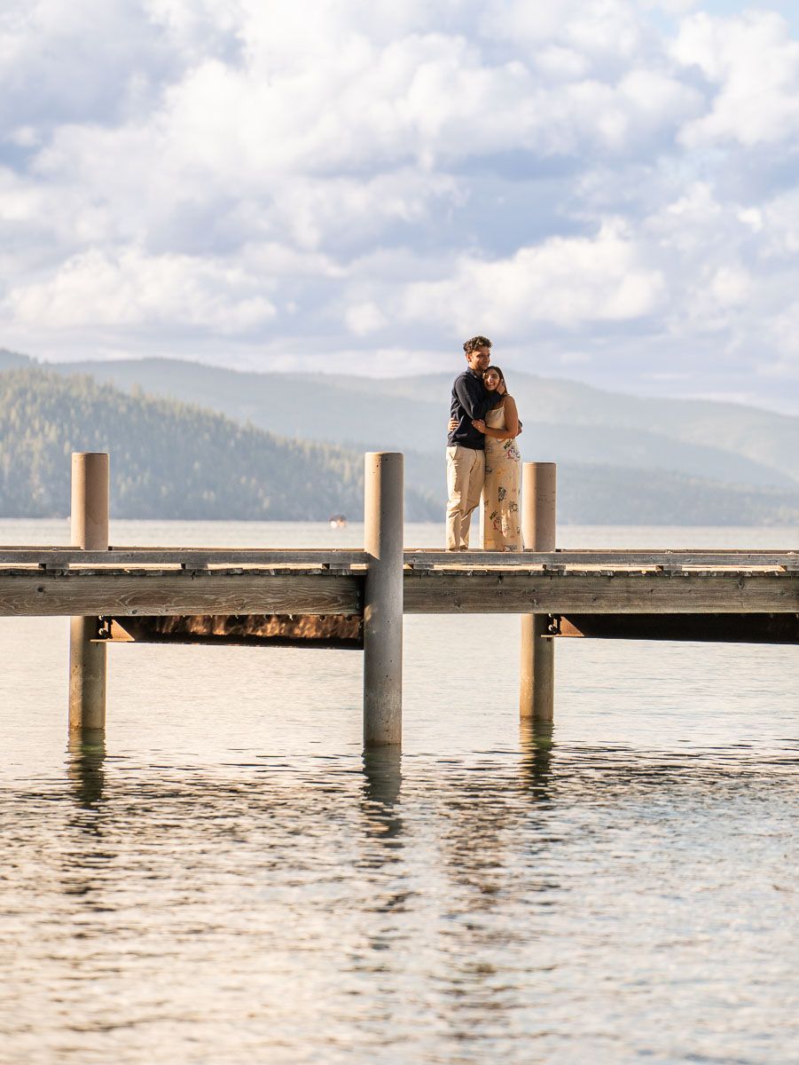 Couple embracing on a wooden dock over calm lake water with mountain and cloudy sky backdrop.