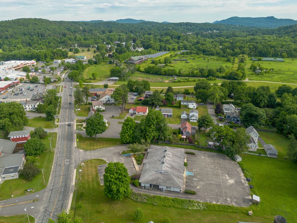 Aerial view of a small town commercial strip with roads, parking lots, green fields, and mountain backdrop.