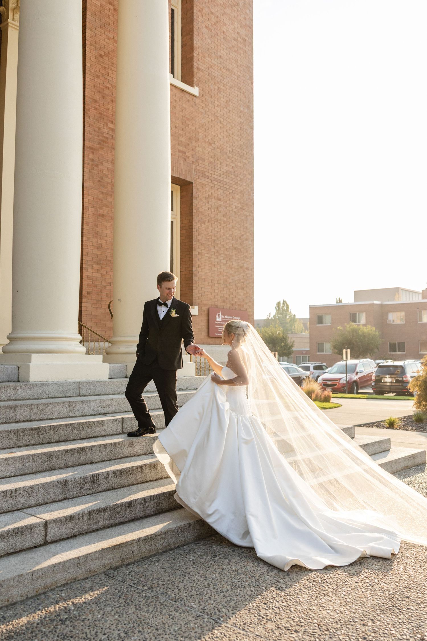 Bride in flowing white gown ascending church steps, groom in black tux extending hand, veil trailing behind.