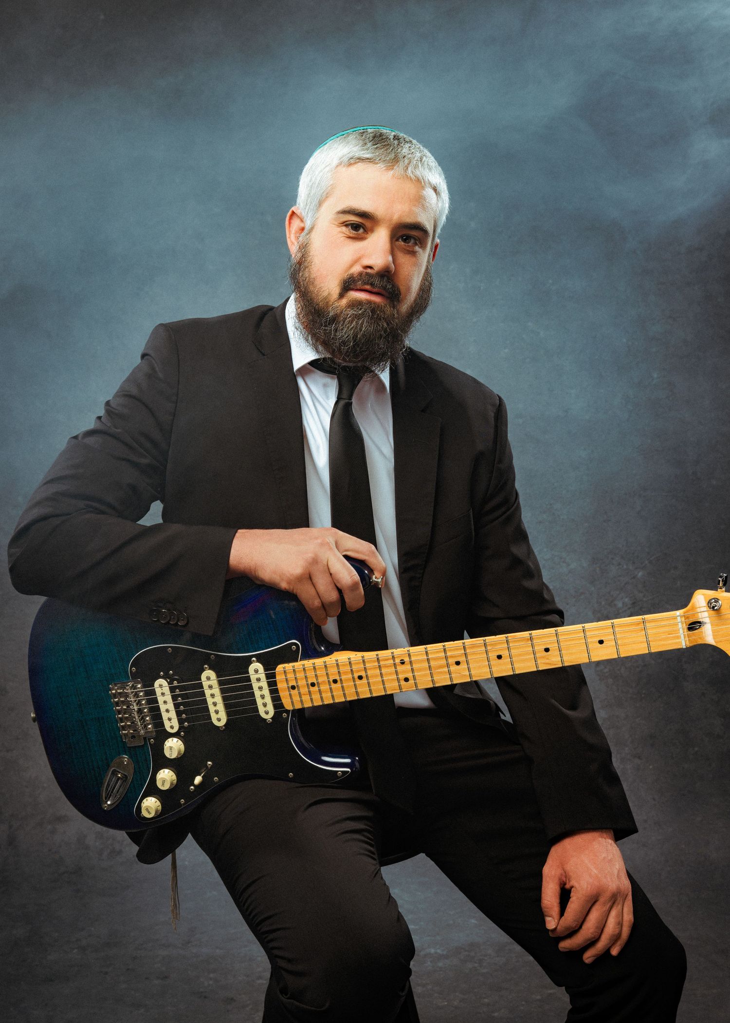 Bearded man with white hair in a black suit holding a blue electric guitar against a smoky gray background.