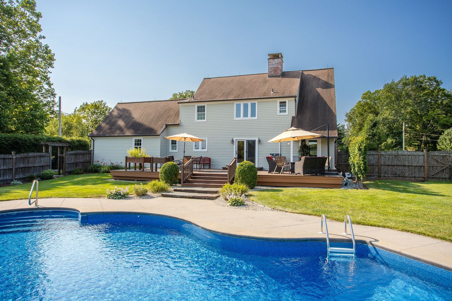 Large suburban home with in-ground pool, wooden deck, patio umbrellas, and lush green lawn on a sunny day.