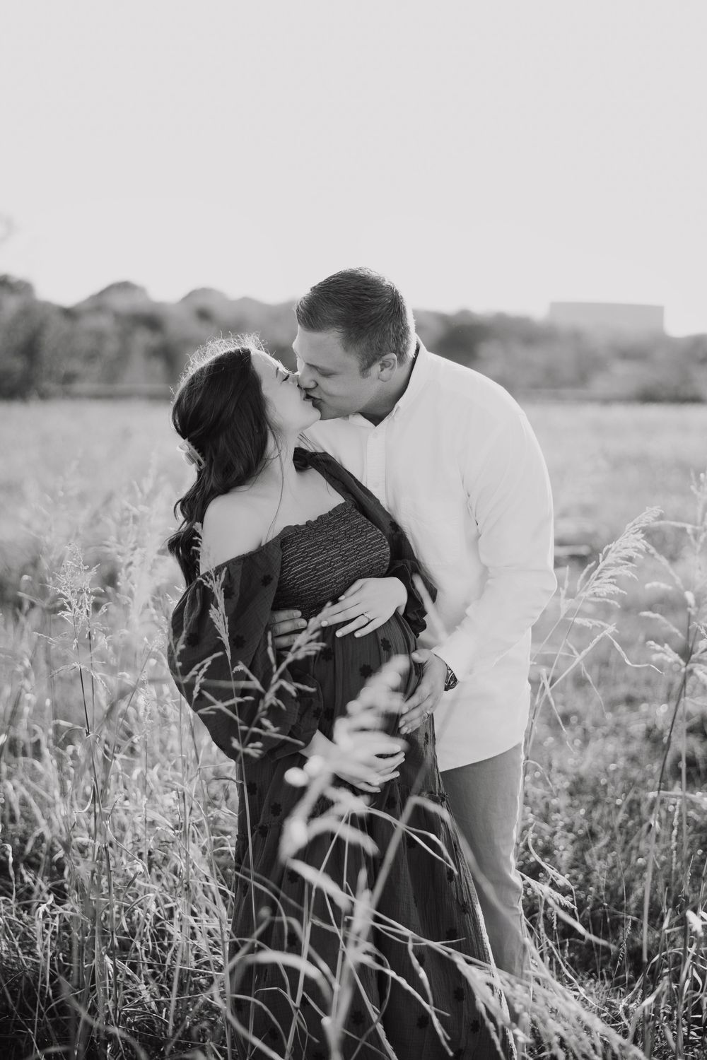 A romantic couple embraces tenderly in a sunlit field of tall grass, captured in black and white photography.