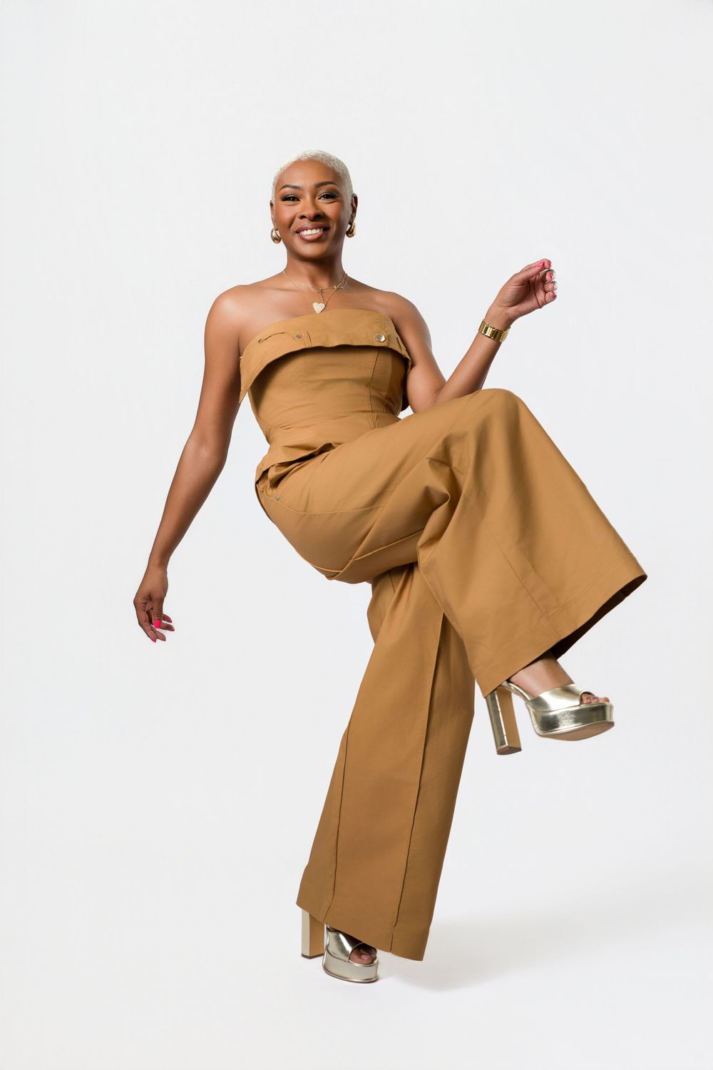 Model wearing a camel-colored strapless jumpsuit with dramatic wide legs and metallic heels against white background.