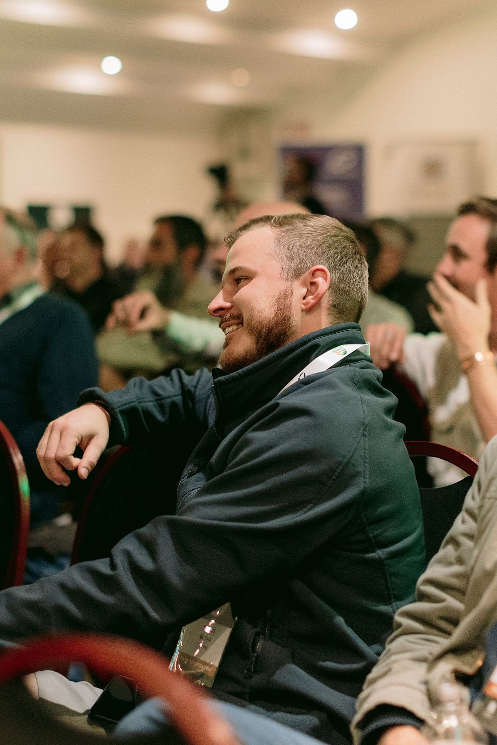 A smiling bearded man in a navy jacket laughs while seated at an indoor conference or event with attendees in background.