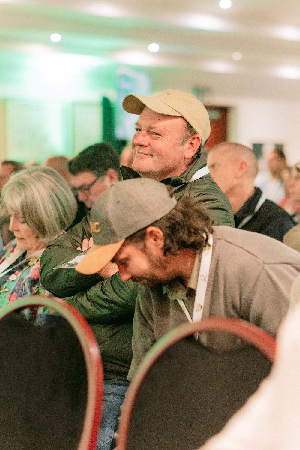 Man in beige cap smiling at a conference event surrounded by seated attendees in a bright indoor venue.